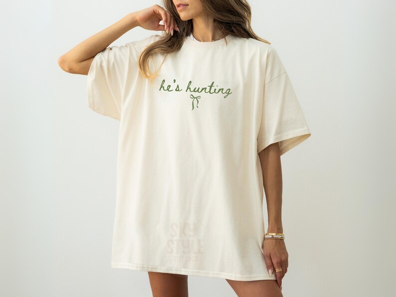 以下が含まれることがあります： クリーム色のオーバーサイズのTシャツ。緑色の筆記体で「he's hurting」と書かれ、リボンが描かれています。半袖でゆったりとしたシルエット。カジュアルなスタイルに最適です。柔らかい素材です。