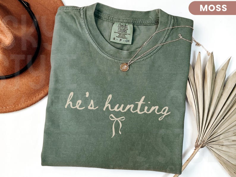 以下が含まれることがあります： モスグリーンのTシャツ。テキスト「he's hunting」は薄いベージュのスクリプト。 金色のネックレスとペンダントがシャツの上にあります。 茶色のカウボーイハットと乾燥したヤシの葉が背景にあります。
