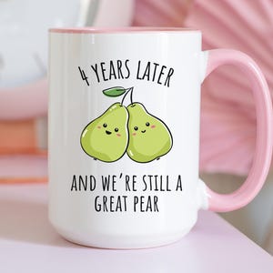 K&ouml;nnte beinhalten: Wei&szlig;er Keramikbecher mit rosa Henkel und Rand. Der Becher zeigt den Text "4 YEARS LATER AND WE'RE STILL A GREAT PEAR" mit einer Illustration von zwei l&auml;chelnden Birnen. Der Becher steht auf einer rosa Oberfl&auml;che.