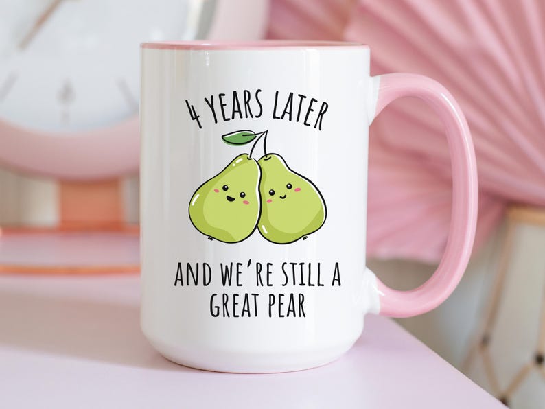 K&ouml;nnte beinhalten: Wei&szlig;er Keramikbecher mit rosa Henkel und Rand. Der Becher zeigt den Text "4 YEARS LATER AND WE'RE STILL A GREAT PEAR" mit einer Illustration von zwei l&auml;chelnden Birnen. Der Becher steht auf einer rosa Oberfl&auml;che.