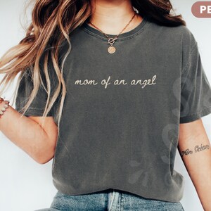 K&ouml;nnte beinhalten: Pfefferfarbenes T-Shirt mit dem gestickten Text "mom of an angel". Das Shirt hat einen Rundhalsausschnitt und kurze &Auml;rmel. Das Model tr&auml;gt eine goldene Halskette und Armb&auml;nder.