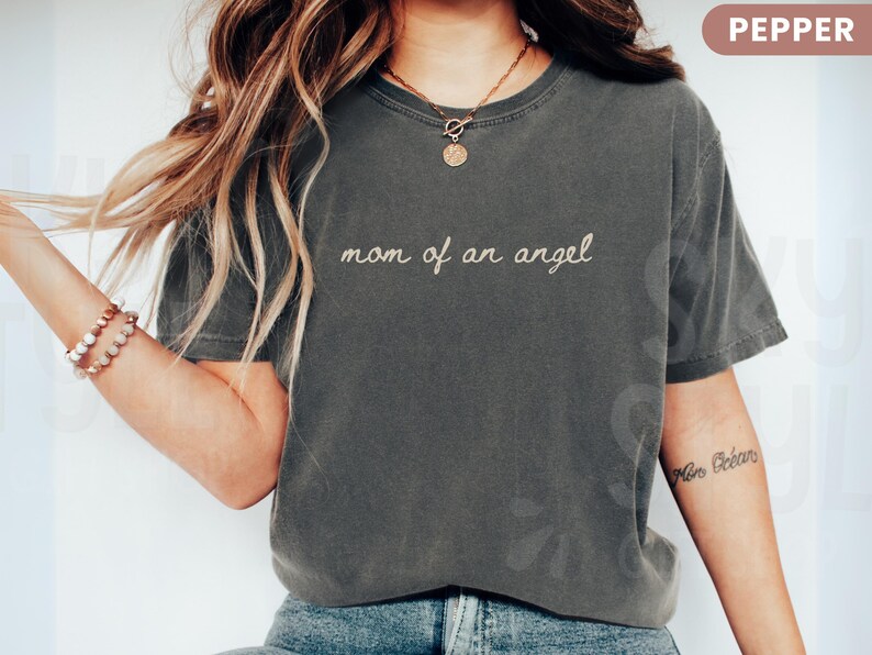 K&ouml;nnte beinhalten: Pfefferfarbenes T-Shirt mit dem gestickten Text "mom of an angel". Das Shirt hat einen Rundhalsausschnitt und kurze &Auml;rmel. Das Model tr&auml;gt eine goldene Halskette und Armb&auml;nder.