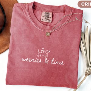 Może przedstawiać: Koszulka w kolorze karmazynowym z napisem "weenies & tinis" i grafiką jamnika. Widoczny jest r&oacute;wnież złoty naszyjnik z wisiorkiem i brązowy kapelusz. Koszulka wykonana jest z miękkiego materiału.