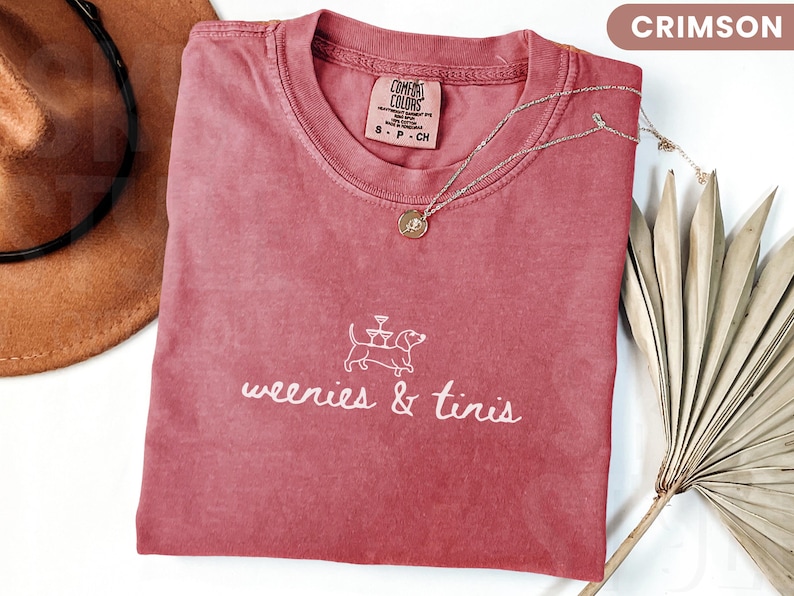 Może przedstawiać: Koszulka w kolorze karmazynowym z napisem "weenies & tinis" i grafiką jamnika. Widoczny jest r&oacute;wnież złoty naszyjnik z wisiorkiem i brązowy kapelusz. Koszulka wykonana jest z miękkiego materiału.