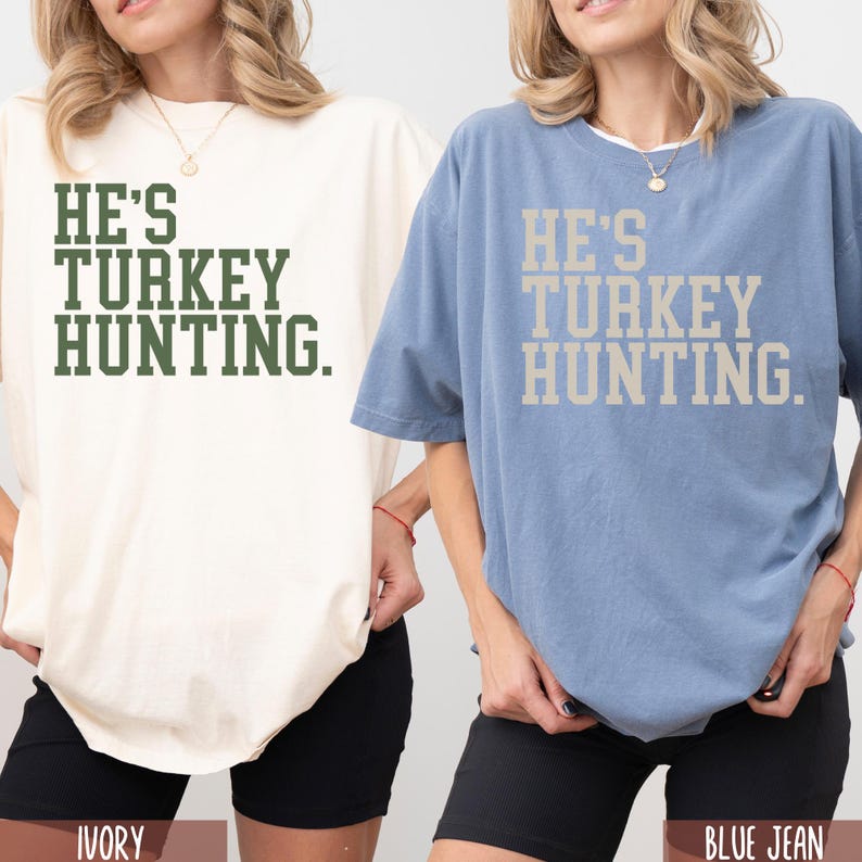 Puede incluir: Dos mujeres con camisetas de gran tama&ntilde;o con el texto "He's Turkey Hunting." La camiseta de la izquierda es de color marfil y la camiseta de la derecha es de color azul vaquero.