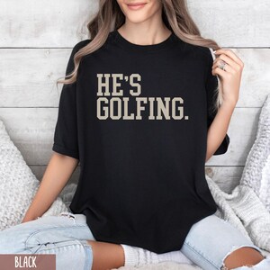 May include: Black t-shirt with the text "HE'S GOLFING." in a beige color.