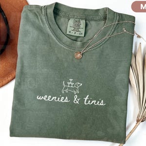 Może przedstawiać: Koszulka w kolorze mchu z napisem "weenies & tinis" i ilustracją jamnika. Złoty naszyjnik z wisiorkiem spoczywa na koszulce. Brązowy kapelusz i suszone liście palmowe znajdują się w tle.