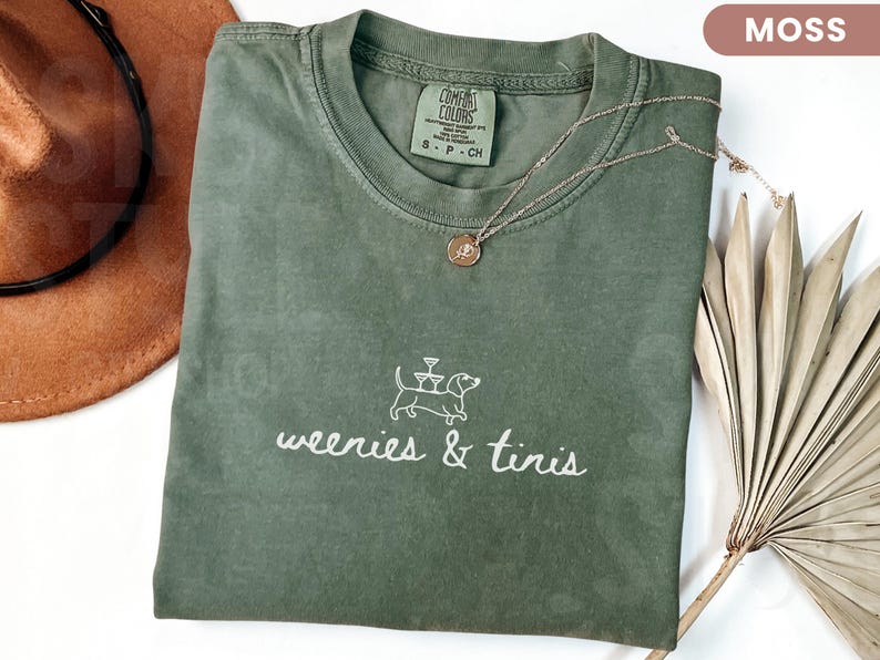 Może przedstawiać: Koszulka w kolorze mchu z napisem "weenies & tinis" i ilustracją jamnika. Złoty naszyjnik z wisiorkiem spoczywa na koszulce. Brązowy kapelusz i suszone liście palmowe znajdują się w tle.