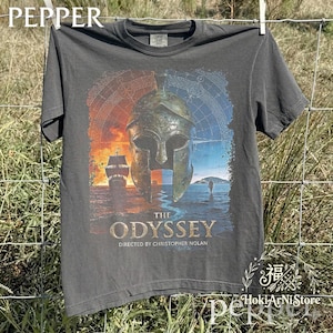 May include: Dark grey t-shirt with a graphic from "The Odyssey" film. The design features a Spartan helmet, a ship, and the text "THE ODYSSEY DIRECTED BY CHRISTOPHER NOLAN". The t-shirt is hanging on a clothesline against a natural background.