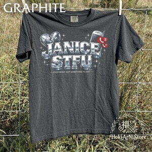 Puede incluir: Una camiseta gris carbón de manga corta con un gráfico de estilo metálico que dice JANICE STFU y EVERYBODY GOT SOMETHING TO SAY. El diseño incluye un icono de corazón roto, un teléfono móvil vintage y una burbuja de notificación roja con un 3.