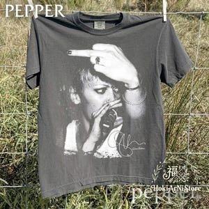 May include: Dark grey t-shirt with a black and white image of a person holding a microphone and giving the middle finger. The word "PEPPER" is printed at the top of the shirt.