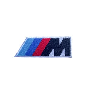 Può includere: Toppa ricamata con il logo BMW M. La toppa ha una forma rettangolare con un bordo bianco. Il logo è composto da strisce blu, blu scuro, rosse e nere e dalla lettera "M".