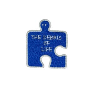 May include: Blue embroidered puzzle piece patch with white stitching and the text "THE DEBRIS OF LIFE".