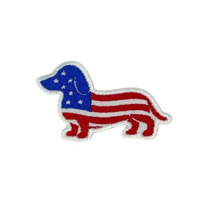 Puede incluir: Parche bordado con un diseño de perro salchicha. La cabeza del perro es azul con estrellas blancas, y el cuerpo es de rayas rojas y blancas, asemejándose a la bandera estadounidense. El parche tiene un borde blanco.