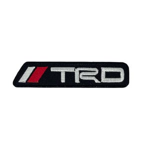 May include: Black embroidered patch featuring the letters "TRD" in white, and a red and white stripe design. The patch has a rectangular shape with rounded corners. Likely for car enthusiasts.