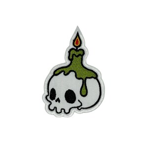 Pode incluir: Patch bordado com uma caveira branca com órbitas pretas e uma vela verde. A vela tem uma chama amarela e vermelha. O patch tem uma borda branca, ideal para roupas ou acessórios.