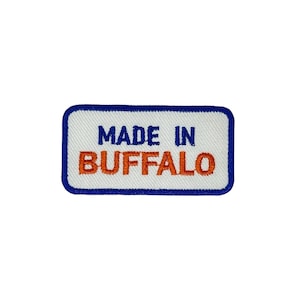 以下が含まれることがあります： 白地の長方形パッチで、青い縁取りがあります。「MADE IN BUFFALO」の文字が青とオレンジの糸で刺繍されています。このパッチは、衣類やアクセサリー用である可能性があります。