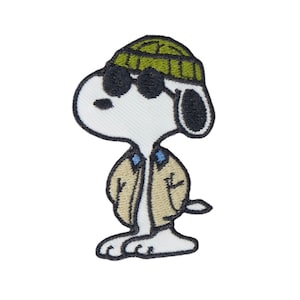 Op de afbeelding: Geborduurde patch met Snoopy met een zonnebril, een groene muts en een beige jas. De patch is wit met zwarte, groene, beige en blauwe accenten. Het ontwerp is een stripfiguur uit de Peanuts-strip.
