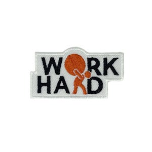 Peut inclure: Écusson brodé avec les mots "WORK HARD" en noir sur fond blanc. Une figurine orange est représentée en train de soulever une grande sphère orange. L'écusson a une forme distinctive avec des bords arrondis.