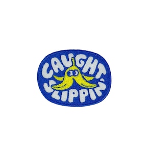 Peut inclure: Un écusson ovale bleu brodé avec le texte "Caught Slippin'" en blanc et une banane jaune avec des yeux au centre.