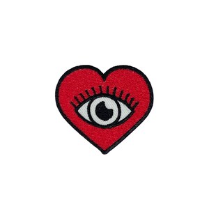 May include: A red heart-shaped patch with a black outline and a white eye design in the center. The eye has a black pupil and black eyelashes. The patch is likely for clothing or accessories.