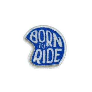Puede incluir: Parche bordado con forma de casco de motocicleta, con las palabras "BORN TO RIDE" en letras blancas sobre fondo azul. El parche tiene un borde blanco con un contorno beige.