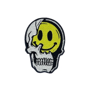 Op de afbeelding: Een witte schedel met een gele smiley die de helft van de schedel bedekt. De schedel is geborduurd met zwarte draad.