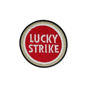 Peut inclure: Écusson rond avec l'inscription "LUCKY STRIKE" en blanc sur fond rouge. Il présente une bordure beige et un bord extérieur noir. Ce patch est probablement destiné à être appliqué sur des vêtements ou des accessoires.