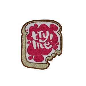 Könnte beinhalten: Ein gestickter Aufnäher in Form einer Scheibe Brot mit dem Text "Try Me!" darin, in Rosa und Weiß.