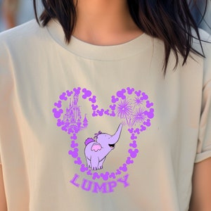 May include: A light beige t-shirt with a purple Mickey Mouse silhouette. Inside the silhouette are purple images of fireworks, a castle, and a purple elephant with the word "Lumpy" underneath.