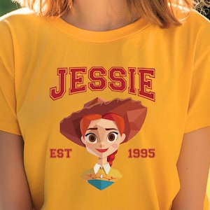 May include: A yellow t-shirt featuring a graphic of a cartoon cowgirl with red hair and a brown hat. The word "JESSIE" is printed above the graphic in red letters, with "EST 1995" below.