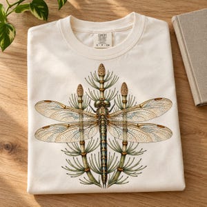 May include: A cream-colored t-shirt featuring a detailed graphic of a dragonfly centered between three horsetail plants. The shirt has a crew neckline and a Comfort Colors brand label printed on the inside of the collar.