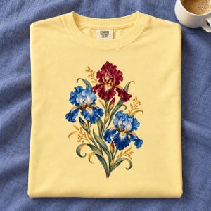 May include: A pale yellow t-shirt featuring a central graphic of three iris flowers, one deep red and two blue, with green stems and golden foliage. The inner neck label displays the text Comfort Colors in black lettering on a white background.