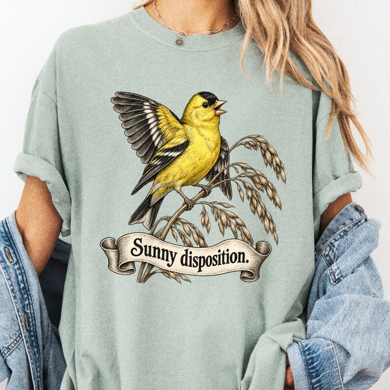 Sage tee close-up on a model with a yellow bird spreading one wing above Sunny Disposition ribbon.