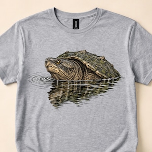 May include: A heather gray short-sleeve t-shirt featuring a detailed graphic illustration of a snapping turtle partially submerged in water. The turtle has a textured, dark olive-green shell and skin, with a clear reflection visible on the water surface.
