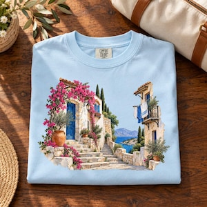 May include: A light blue t-shirt featuring a graphic print of a Mediterranean coastal scene with stone steps, a blue door, pink bougainvillea flowers, and hanging laundry. The inner neck label displays the text Comfort Colors.