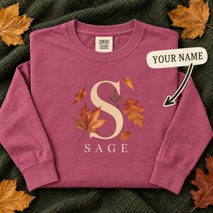 May include: A long-sleeve crewneck sweatshirt in a muted berry color. The front features a large cream-colored letter S surrounded by printed autumn leaves in shades of orange and brown. Below the letter, the word SAGE is printed in cream text.