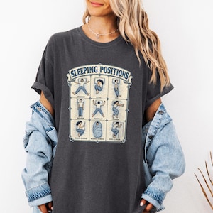May include: A charcoal gray short-sleeve t-shirt featuring a graphic print titled SLEEPING POSITIONS. The design displays a grid of nine illustrations showing various human figures in different sleeping postures with labels underneath each one.