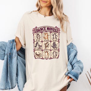 May include: A cream-colored t-shirt featuring a graphic print titled DANCE MOVES, displaying nine illustrations of cats performing various dance steps, including The Twist, The Sprinkler, The Robot, The Shopping Cart, The Running Man, The Lawn Mower, The Worm, Disco Point, and The Floss.