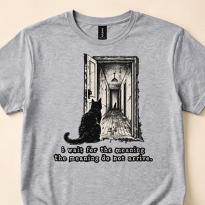 May include: A heather gray t-shirt featuring a black ink-style graphic of a cat sitting in a doorway looking down a hallway toward another cat. The text below the graphic reads, i wait for the meaning the meaning do not arrive.