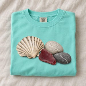 May include: A turquoise crew neck t-shirt with a Comfort Colors label at the inner neckline. The front features a graphic print of a large tan seashell, a speckled light gray stone, a dark gray stone, and a smooth deep red sea glass piece.