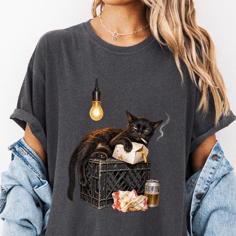 Close view of a charcoal tee with a lounging black cat, food wrappers and hanging bulb.