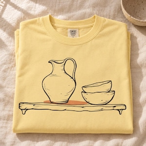May include: A pale yellow t-shirt featuring a black line art graphic of a pitcher and two stacked bowls resting on a wooden shelf. The shirt has a Comfort Colors brand label at the inner neckline with the size L clearly printed on the tag.