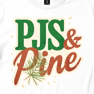 May include: A white t-shirt featuring a graphic design with the text PJS & Pine. The word PJS is in green, and the word Pine is in a reddish-brown script font. A green pine needle sprig is positioned over the letter P in the word Pine.