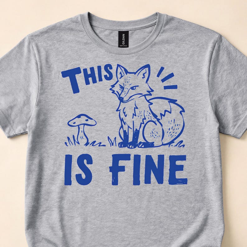 Close view of a gray T-shirt with a blue fox beside a mushroom and This Is Fine lettering.