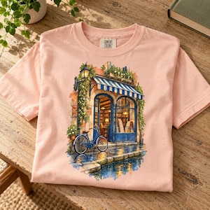 May include: A light peach-colored t-shirt featuring a graphic print of a bookstore with a blue striped awning, a blue bicycle parked in front, and a lamppost. The inner neck label displays the text Comfort Colors in black lettering.