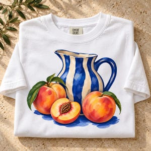 May include: A white short-sleeve t-shirt with a Comfort Colors label at the neck. The graphic features a blue and white striped pitcher surrounded by three peaches, one of which is sliced in half to reveal the pit, all with green leaves.