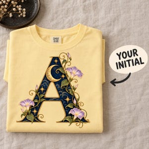 May include: A pale yellow t-shirt featuring a large letter A graphic. The letter is filled with a dark blue night sky pattern, a crescent moon, and stars, surrounded by purple morning glory flowers and green vines. The neck label reads Comfort Colors.
