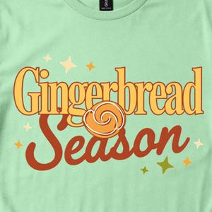 May include: A light green t-shirt featuring the text Gingerbread Season in a mix of yellow serif and brown script fonts. A small, circular, orange-brown cookie graphic with white icing swirls sits in the center, surrounded by small yellow stars.