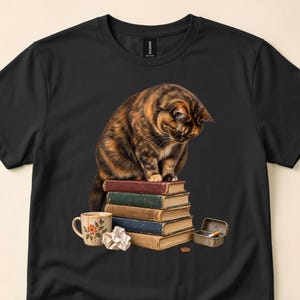 May include: A black t-shirt featuring a graphic of a brown and black tabby cat sitting on a stack of five vintage books. Beside the books is a white ceramic mug with a floral pattern, a crumpled piece of paper, a small coin, and an open metal tin.